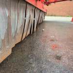 Rusty metal skirt with curved vertical blades along the bottom edge of a large red industrial machine, on a wet pavement.