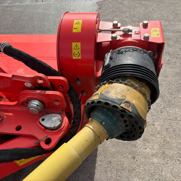 Close-up of red hydraulic machinery with yellow warning stickers and a thick yellow hose on a concrete surface.