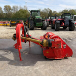 Red agricultural implement in the foreground with a row of tractors and farming equipment on a gravel lot.