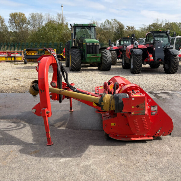 Red agricultural implement in the foreground with a row of tractors and farming equipment on a gravel lot.