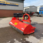 Red agricultural mower in the foreground outside a brick building with a New Holland sign reading 'Agriculture Russells' on blue and yellow panels.