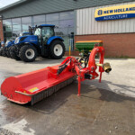 Blue New Holland tractor parked outside an agriculture dealership, with a red farming implement in the foreground and a large glass showroom behind it.