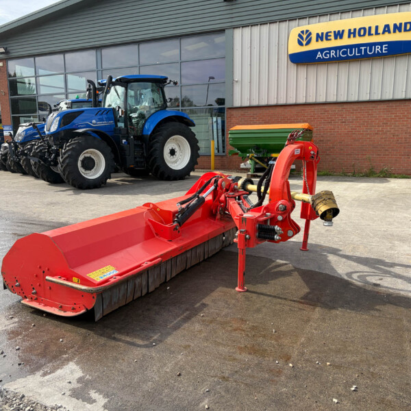 Blue New Holland tractor parked outside an agriculture dealership, with a red farming implement in the foreground and a large glass showroom behind it.