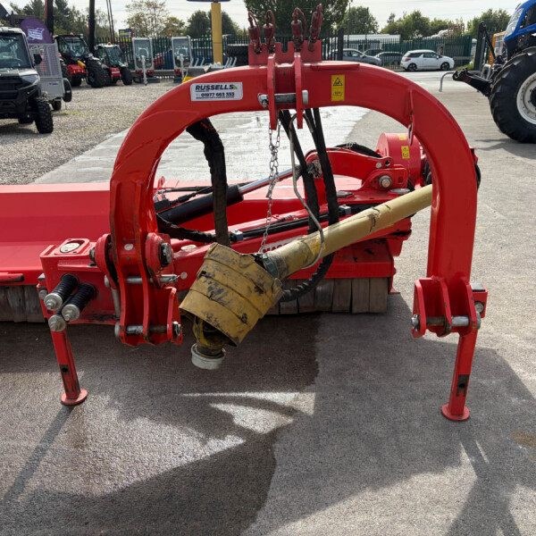 Red farming implement with hydraulic hoses and metal frame parked in a dealership lot; other tractors visible in the background