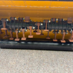 Close-up of a rusted rotating shaft with curved blades mounted in a yellow machine housing, showing bolted joints and wear marks on the metal components.