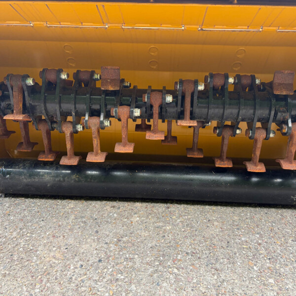 Close-up of a rusted rotating shaft with curved blades mounted in a yellow machine housing, showing bolted joints and wear marks on the metal components.