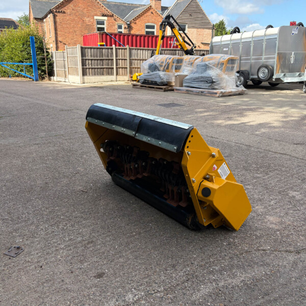 Yellow construction roller in foreground with wrapped equipment on pallets and a silver trailer, in an industrial yard with brick buildings in the background.