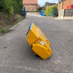 Overturned yellow road safety barrier on a paved street, with blue gates and brick buildings in the background outside a construction area.