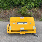 Front view of a bright yellow machine on a gravel surface with a dense green hedge behind it.