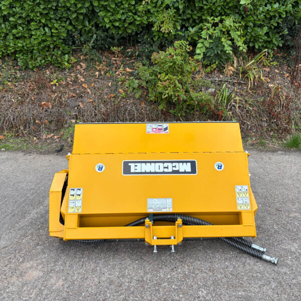 Front view of a bright yellow machine on a gravel surface with a dense green hedge behind it.