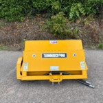 Yellow industrial machinery sits on a paved surface in front of a dense green hedge, viewed from above.