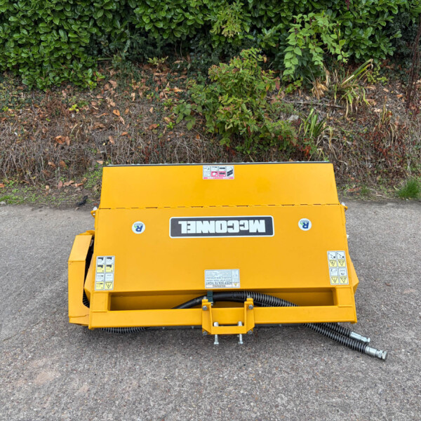 Yellow industrial machinery sits on a paved surface in front of a dense green hedge, viewed from above.