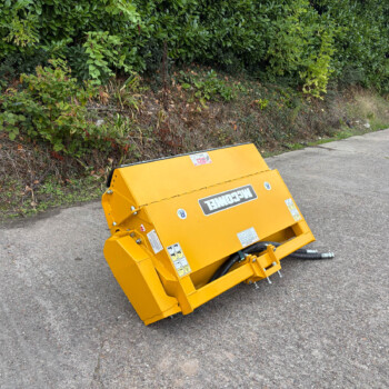 Yellow construction/landscaping machine tipped on a paved road beside a hedge. Far right shows a blue gate and green sign.