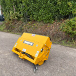 Yellow industrial machine with hoses attached sits on a paved street beside a hedge and a parked red car, near a building with a refuse bin area in the background.