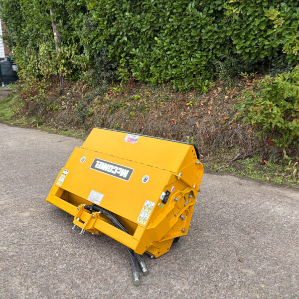 Yellow industrial machine with hoses attached sits on a paved street beside a hedge and a parked red car, near a building with a refuse bin area in the background.