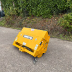 Bright yellow industrial scarifier attachment on a paved driveway beside a tall green hedge; a red car and a building are in the background.