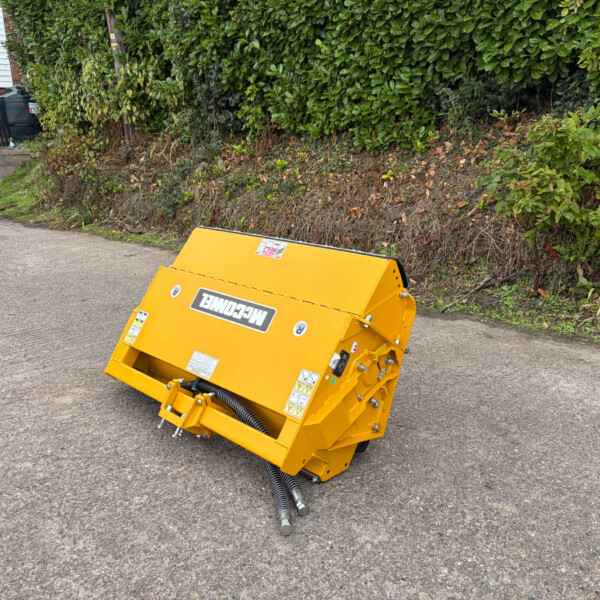 Bright yellow industrial scarifier attachment on a paved driveway beside a tall green hedge; a red car and a building are in the background.