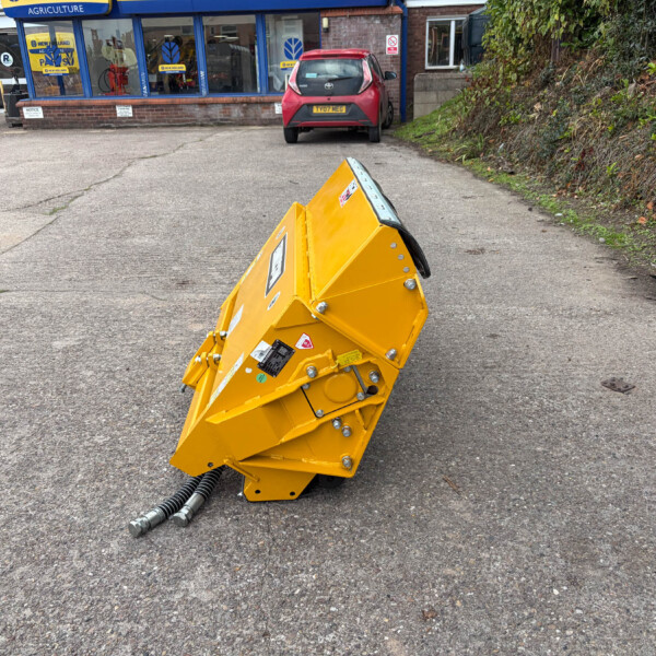 Yellow industrial machine with hoses on a cracked asphalt lot in front of a blue-framed service shop; a man stands to the left using his phone.