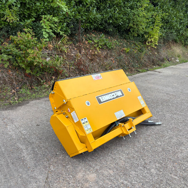 Yellow construction/landscaping machine tipped on a paved road beside a hedge. Far right shows a blue gate and green sign.