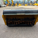 Close-up of a yellow soil roller with a black top and metal bolts, in a yard of farming equipment and tractors in the background.