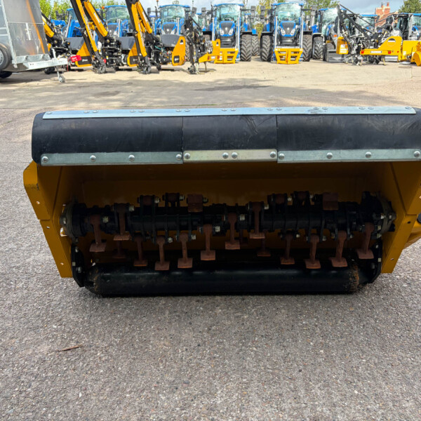 Close-up of a yellow soil roller with a black top and metal bolts, in a yard of farming equipment and tractors in the background.