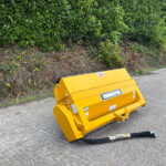 Yellow industrial lawn/yard waste machine resting on a paved road beside a dense hedge, with hoses on the ground nearby.