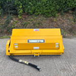 Bright yellow industrial barrier on a paved surface with a black hose in front and a green hedge behind it.