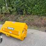Bright yellow construction machine/attachment on a paved surface with a dense green hedge in the background.