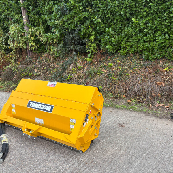 Bright yellow construction machine/attachment on a paved surface with a dense green hedge in the background.