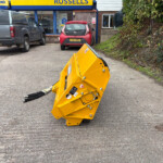 Yellow industrial machinery tipped over in a parking lot in front of a blue-and-yellow New Holland Agriculture shop with cars parked nearby.