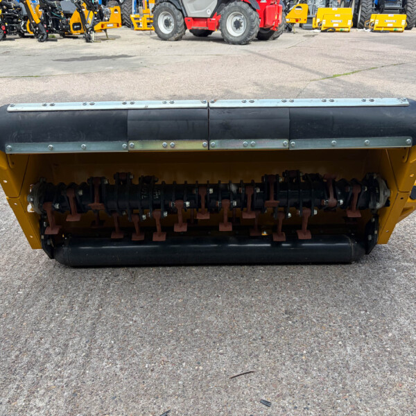 Yellow road roller in the foreground with a large steel drum, parked in an equipment yard with tractors in the background.