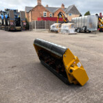 Industrial yard scene with a yellow piece of machinery in the foreground and a trailer carrying blue tractors in the background.