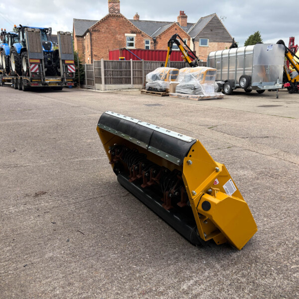 Industrial yard scene with a yellow piece of machinery in the foreground and a trailer carrying blue tractors in the background.