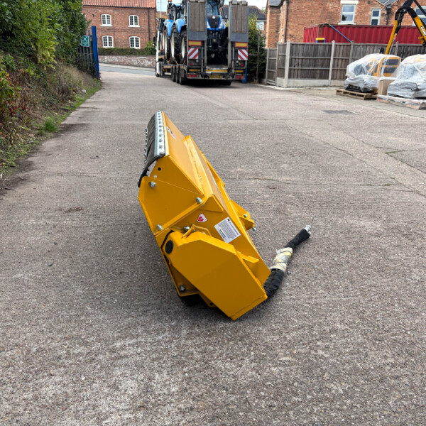 Construction site street with yellow machinery part tipped over on asphalt foreground; trucks and wrapped equipment behind a fence in a residential area