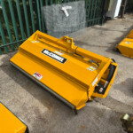 Close-up of a yellow McConnel mower attachment lying on a concrete surface beside a green fence.