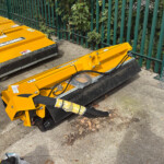 Two large yellow construction machines with black cutting blades sit on a paved surface beside a green metal fence, with leaves scattered nearby.