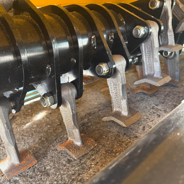 Close-up of a metal crankshaft and curved cutting blades with bolts on a rusted agricultural implement edge.