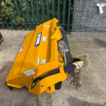 Yellow industrial bucket attachment on a concrete surface beside a green metal fence and stacked wire mesh panels.