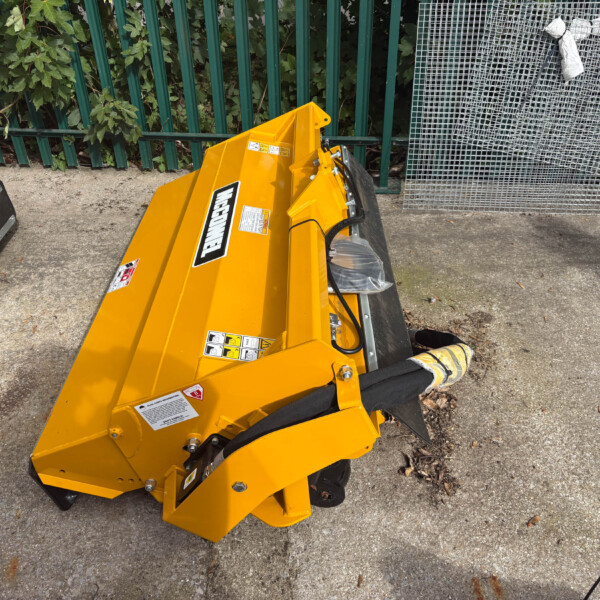 Yellow industrial bucket attachment on a concrete surface beside a green metal fence and stacked wire mesh panels.
