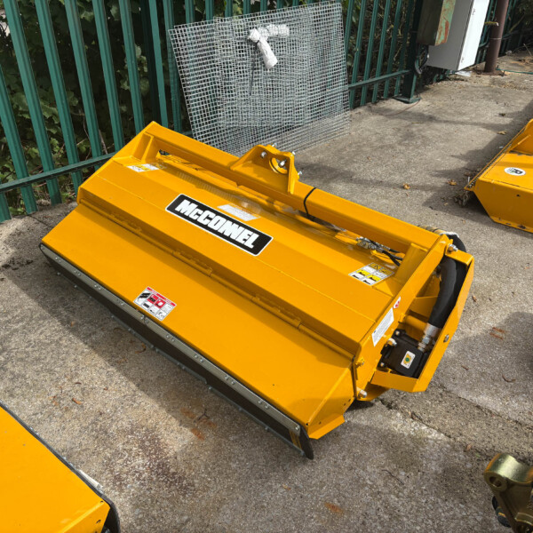 Yellow McConnel mower attachment resting on a concrete surface beside a green metal fence, with a wire mesh panel leaning in the background