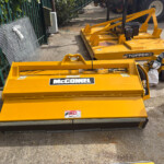 Yellow McConnel Topper 9 mower attachment on a paved surface beside a green fence and scattered equipment.