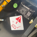 Close-up of a black surface with a green 'Passed Inspection' sticker, a metal identification plate, and a red warning card nearby on a yellow vehicle part.