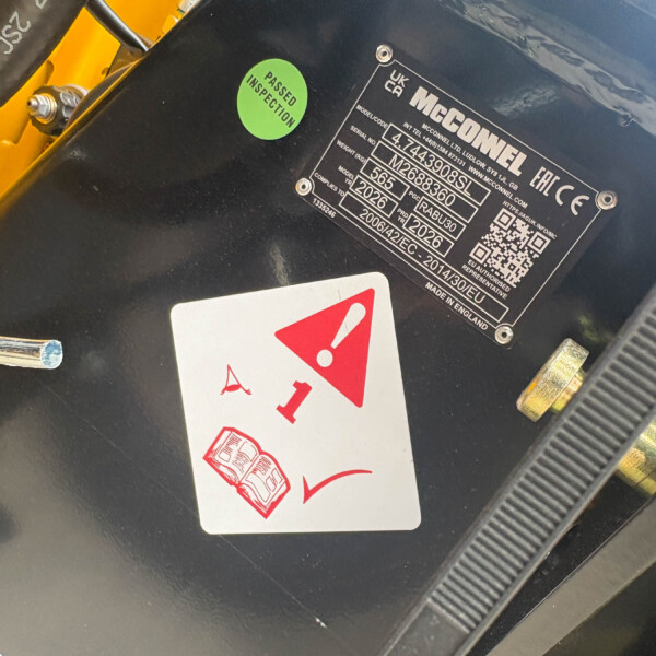 Close-up of a black surface with a green 'Passed Inspection' sticker, a metal identification plate, and a red warning card nearby on a yellow vehicle part.