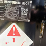 Close-up of an industrial nameplate with model and serial numbers on a dark surface, plus warning triangle decals nearby, hinting at caution around the equipment.