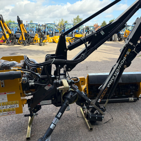 Close-up of a yellow heavy machine with hydraulic arms and hoses in the foreground, row of blue tractors visible in the background, outdoor equipment lot.