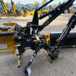 Close-up of a yellow construction attachment with hydraulic hoses, safety decals, and metal arms, in a lot with blue tractors in the background