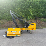 Yellow McConnel stump grinder attachment on a trailer, parked on a paved surface with a green hedge in the background.