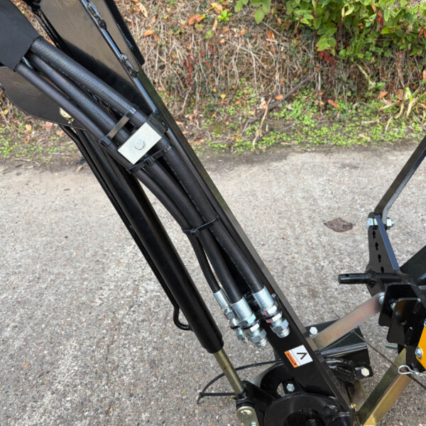 Close-up of hydraulic hoses and metal support frame on a black industrial machine outdoors on gravel, with greenery in the background