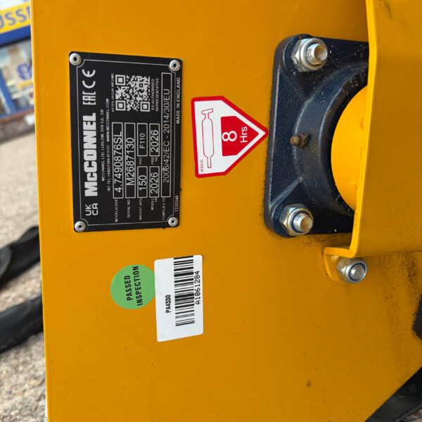 Close-up of a yellow industrial machine side with a metal identification plate, a red 8 hrs safety sticker, and inspection barcode labels.
