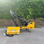 Yellow McConnel agricultural implement with hydraulic arms resting on pavement in front of a green hedge.